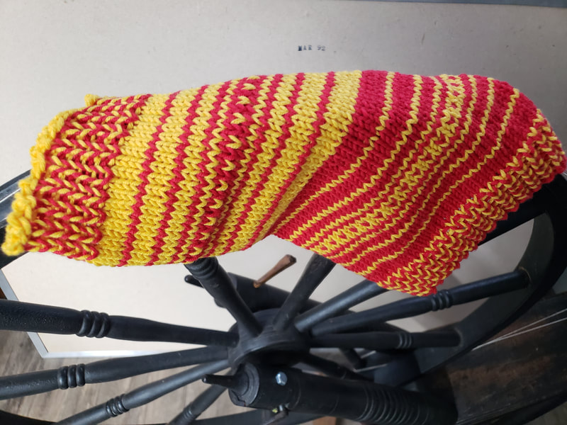 A red and yellow striped knitted cowl draped over the wheel of a spinning wheel. 